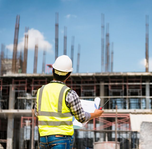 Ein Bauingenieur mit Schutzhelm und Warnweste betrachtet Baupläne vor einer im Bau befindlichen mehrstöckigen Gebäudestruktur unter blauem Himmel.